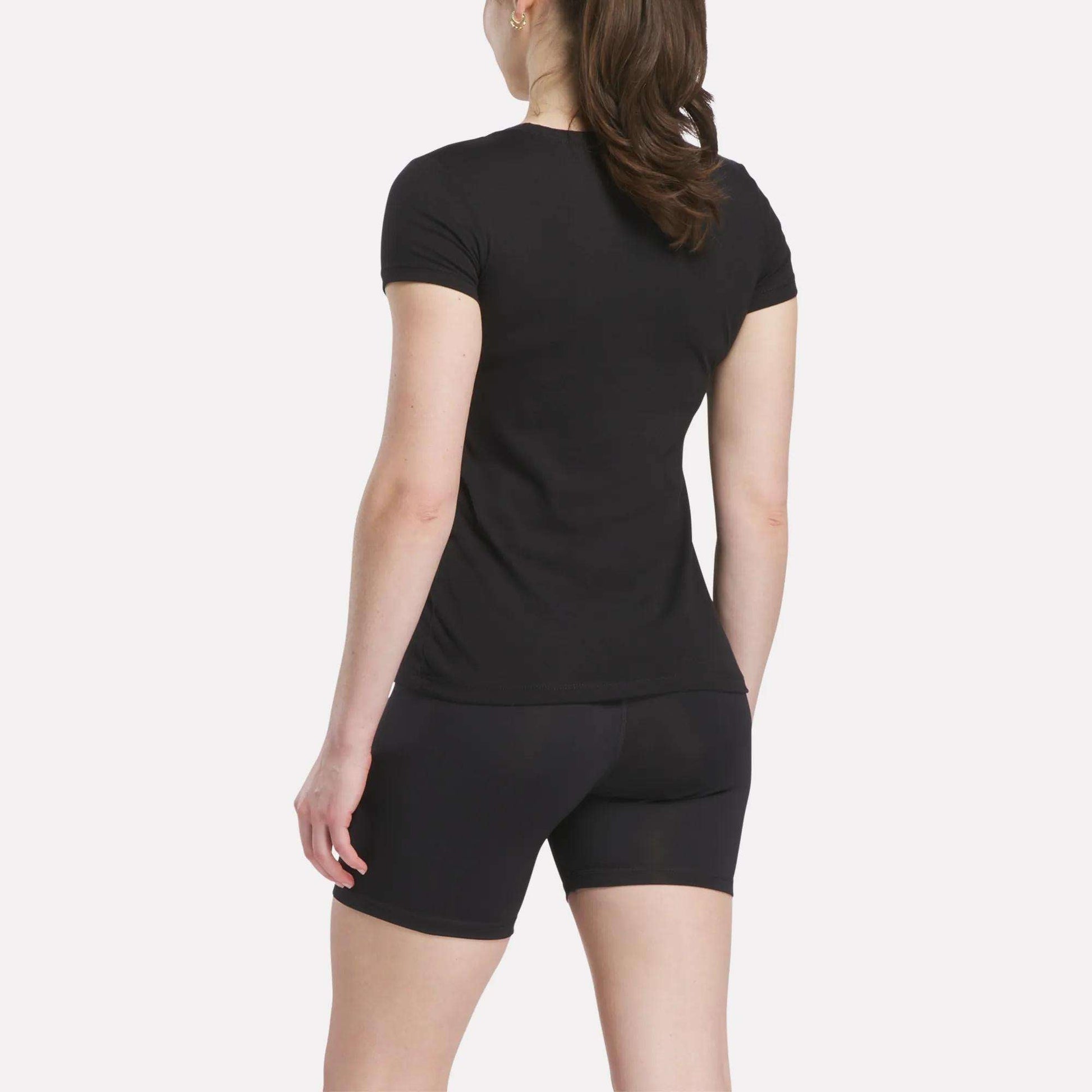 A person with long brown hair in a ponytail is standing and facing away, wearing a Reebok Women's T-Shirt and black shorts against a plain white background.