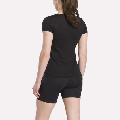 A person with long brown hair in a ponytail is standing and facing away, wearing a Reebok Women's T-Shirt and black shorts against a plain white background.