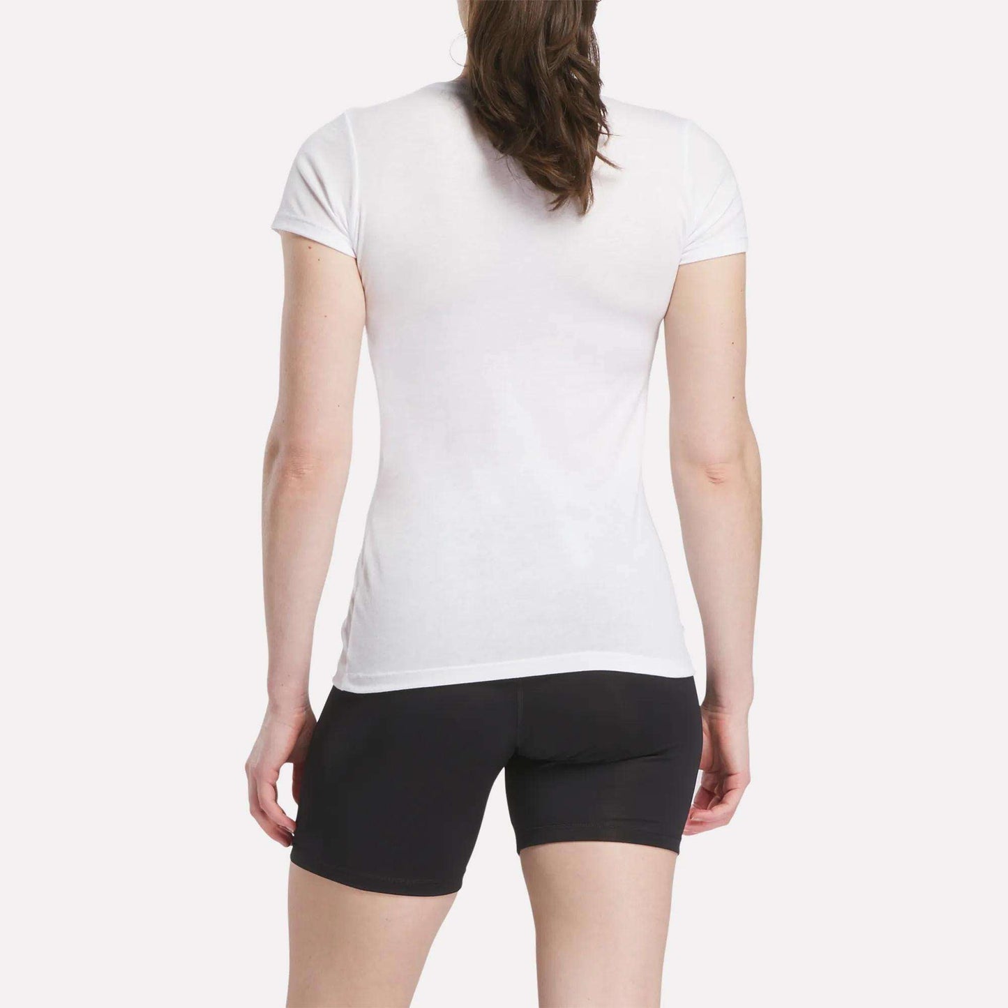 A person with long brown hair is seen from the back, wearing a fitted Women's Reebok T-Shirt and black shorts, standing against a plain white background.