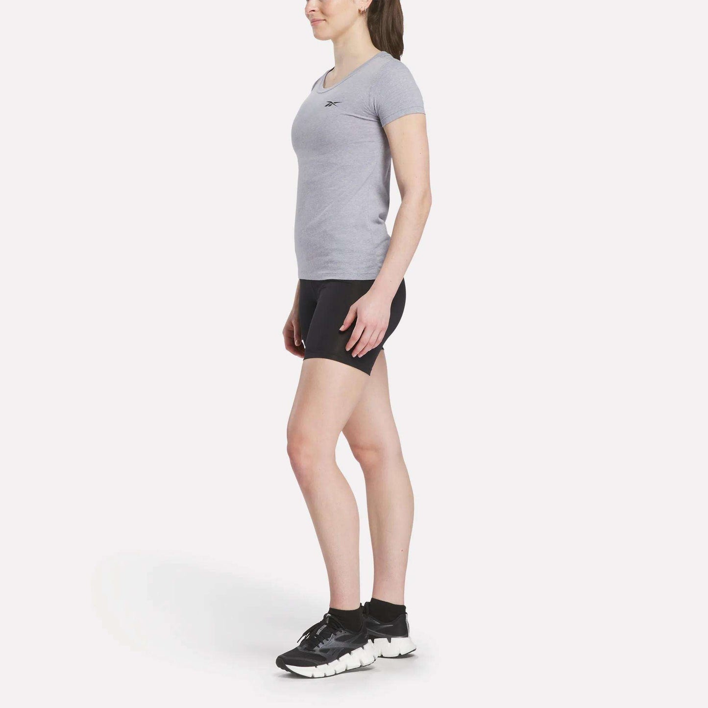 A woman poses in profile against a white background, wearing a fitted gray Women's Reebok T-Shirt, black shorts, black socks, and black athletic shoes.
