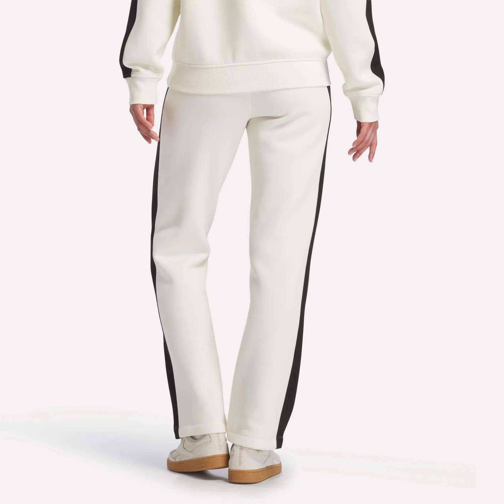 A person is seen from the back standing against a plain background, wearing Reebok Women's Fleece Straight Leg Pants in white with black side stripes, a matching white sweatshirt, and white sneakers.