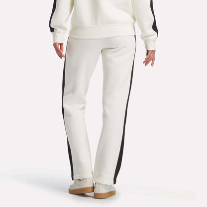 A person is seen from the back standing against a plain background, wearing Reebok Women's Fleece Straight Leg Pants in white with black side stripes, a matching white sweatshirt, and white sneakers.