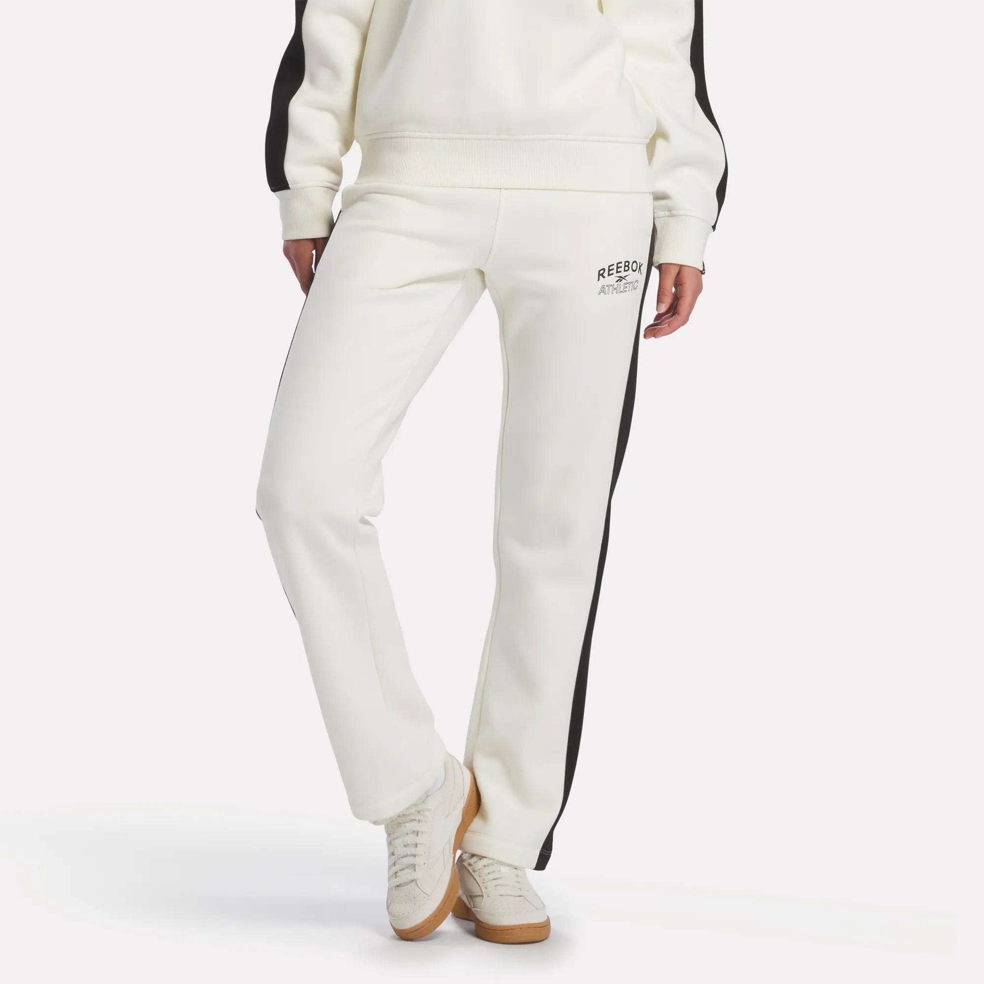 A person is wearing the Reebok Women's Fleece Straight Leg Pants in chalk, featuring black side stripes and a small logo on the upper thigh, paired with white sneakers. Their hands are at their sides against a plain white background.
