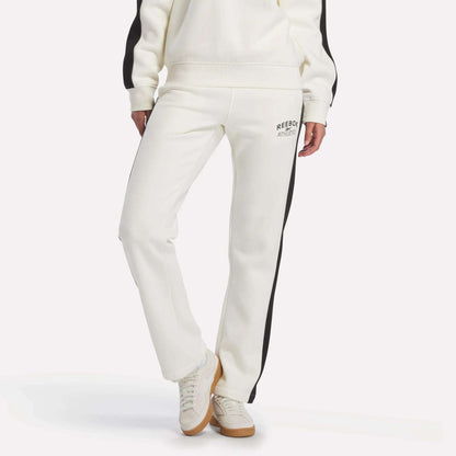 A person is wearing the Reebok Women's Fleece Straight Leg Pants in chalk, featuring black side stripes and a small logo on the upper thigh, paired with white sneakers. Their hands are at their sides against a plain white background.