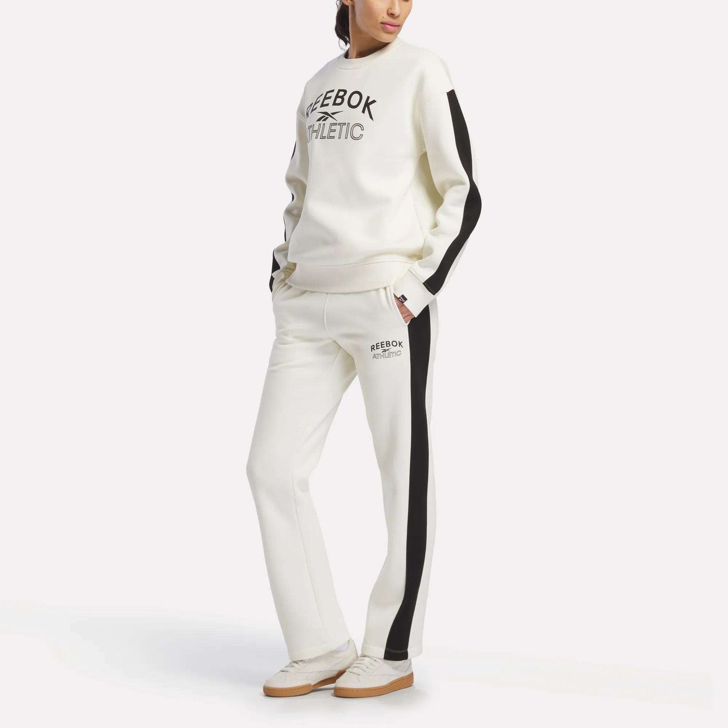 A woman models Reebok Women’s Fleece Straight Leg Pants, featuring black side stripes, paired with a coordinating white sweatshirt; she stands against a plain white background with her hands in her pockets.