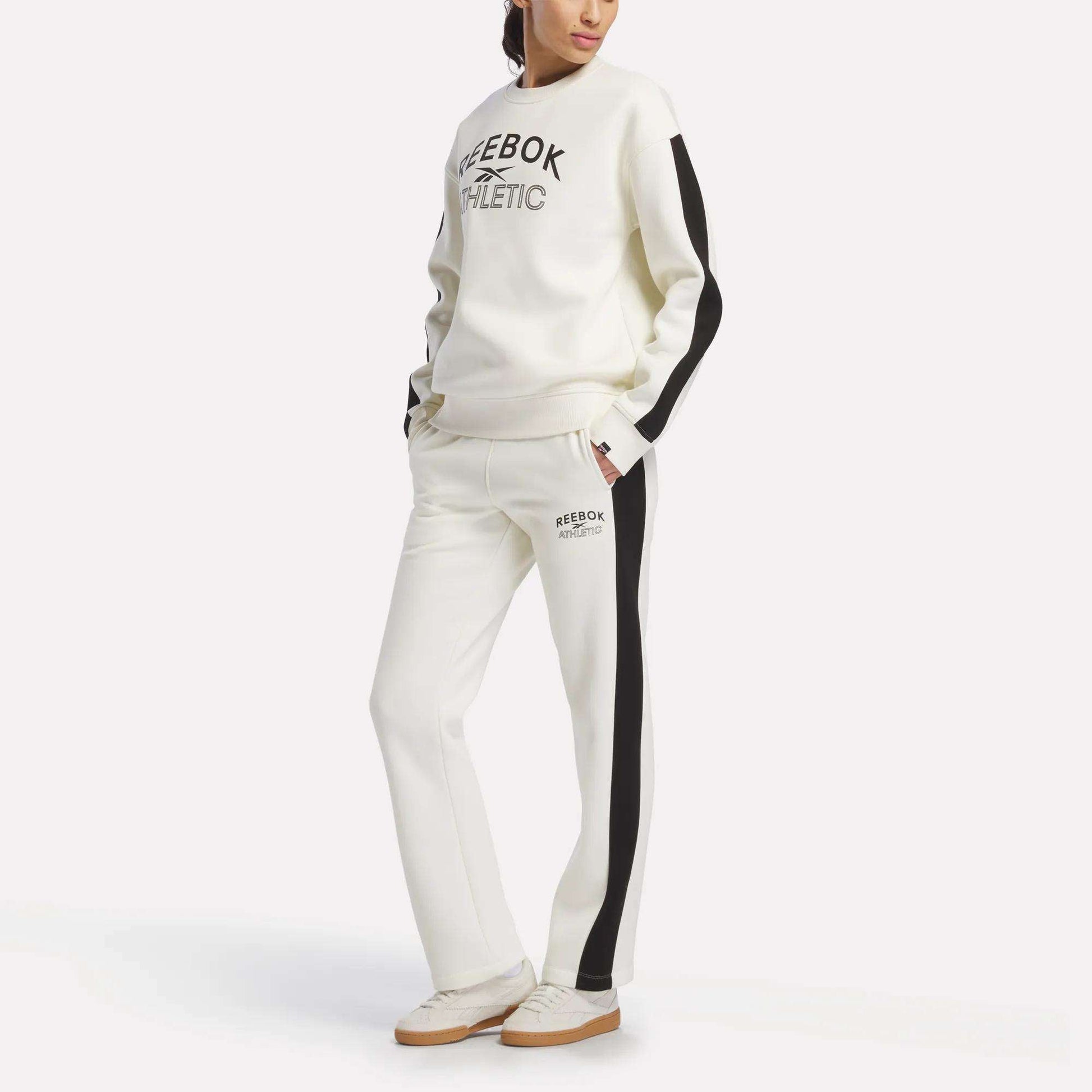 A woman models Reebok Women’s Fleece Straight Leg Pants, featuring black side stripes, paired with a coordinating white sweatshirt; she stands against a plain white background with her hands in her pockets.