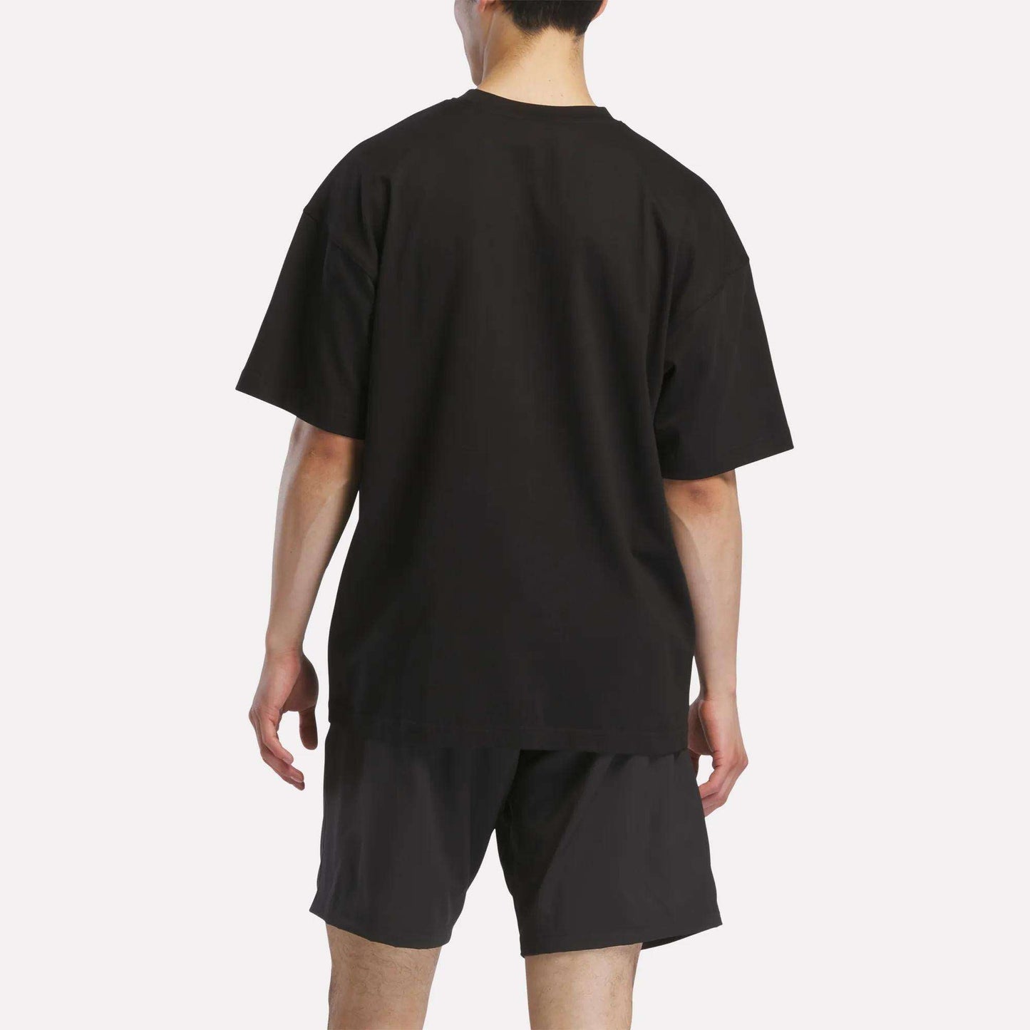 A person faces away from the camera, wearing a loose-fitting black Reebok Men's Basketball T-Shirt and black shorts against a plain white background.