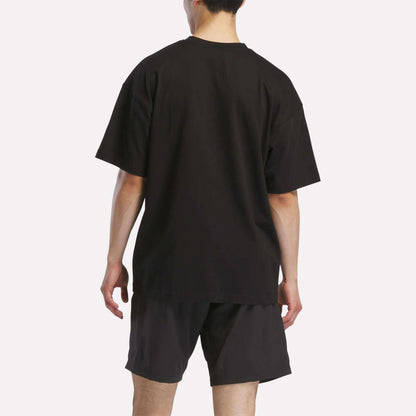 A person faces away from the camera, wearing a loose-fitting black Reebok Men's Basketball T-Shirt and black shorts against a plain white background.