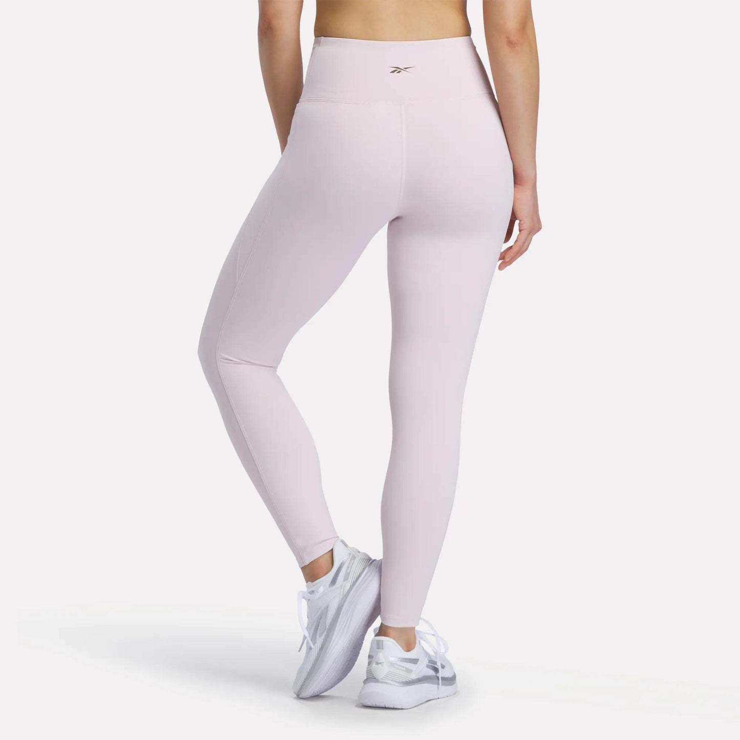 A person is wearing Reebok Women's High-Rise Leggings in light pink, which feature RBK-DRY technology, along with white athletic shoes, standing with their back to the camera against a plain white background.