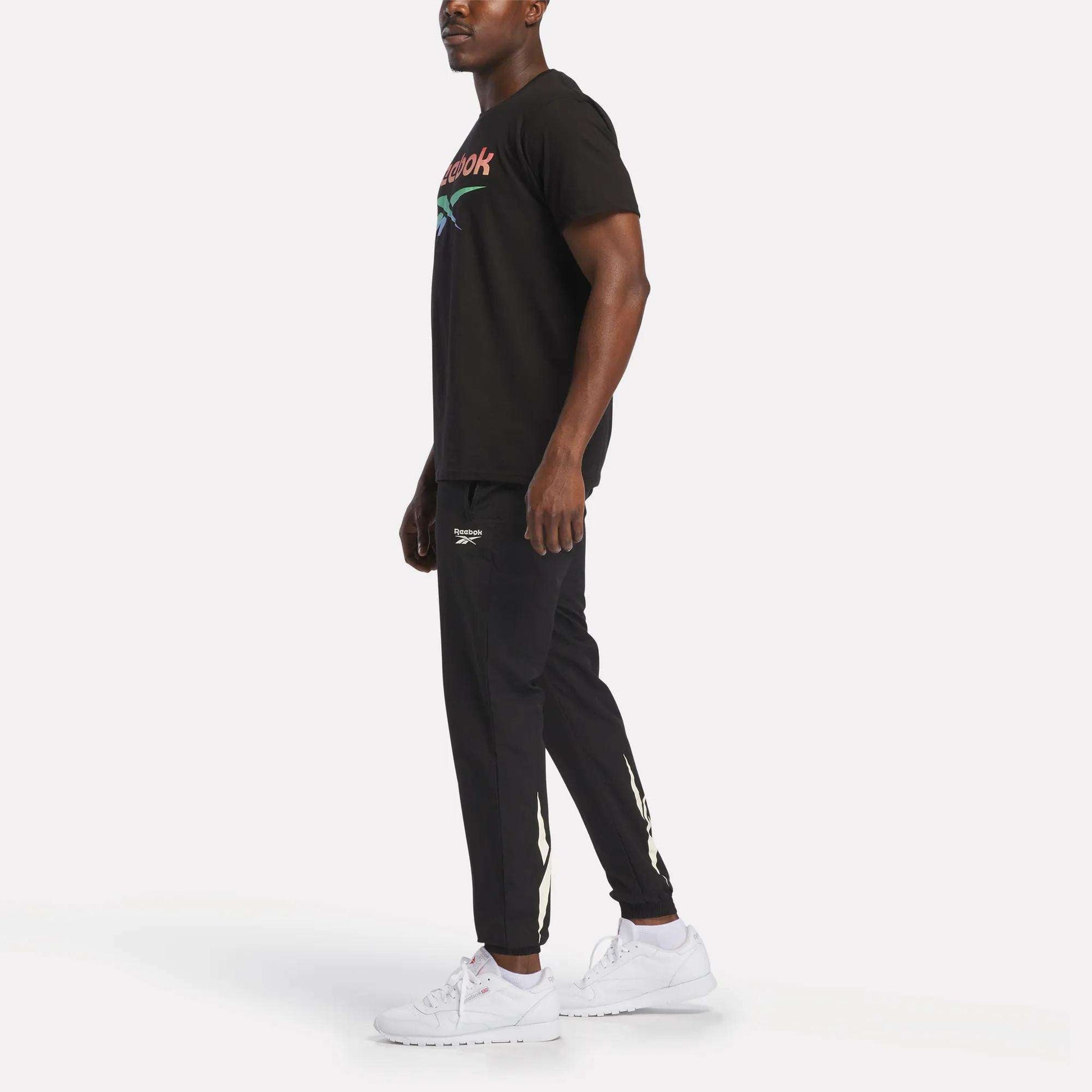 A man stands slightly sideways, wearing Reebok's Men's Vector Graphic Rainbow T-Shirt in black, paired with black joggers with white logo details and white sneakers, against a plain white background.