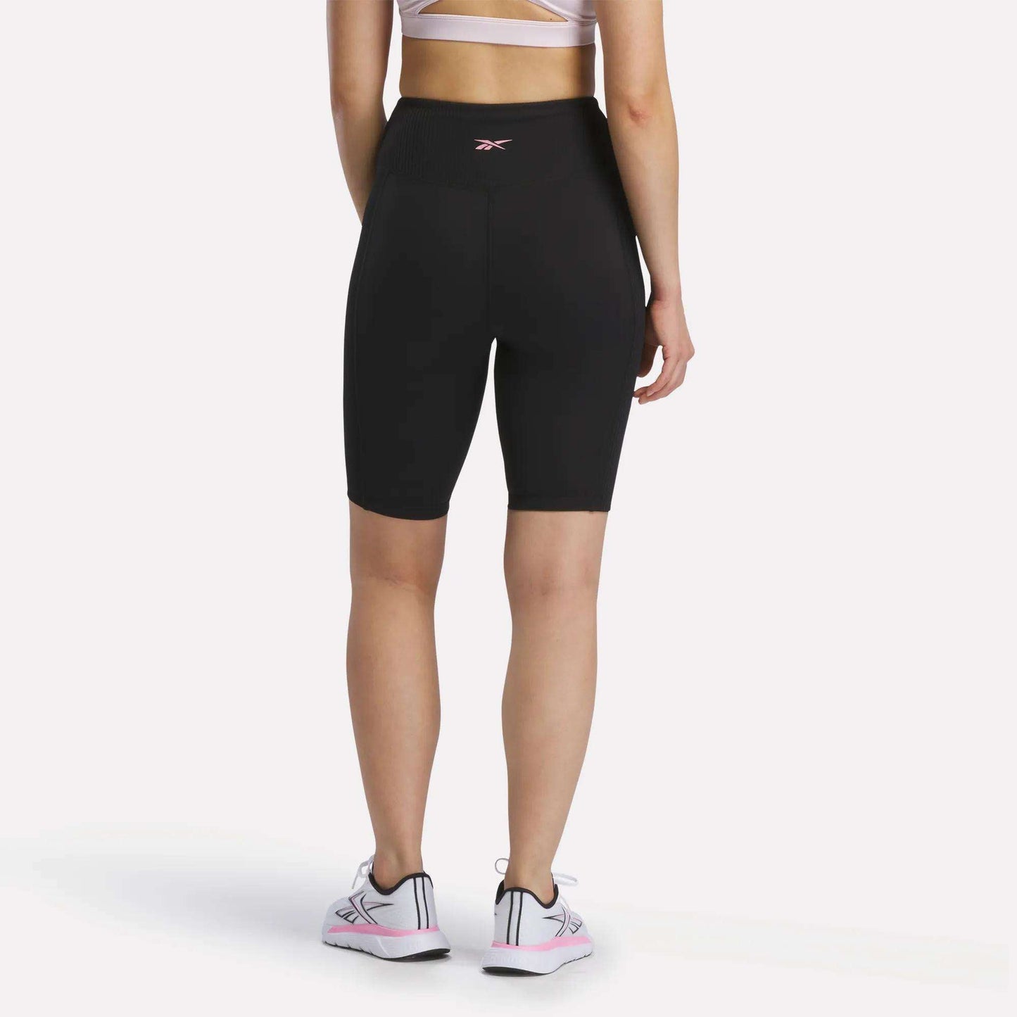 A person stands facing away against a plain white background, wearing a white sports bra, Reebok Women's Peached Biker Shorts 9" in black made from peached fabric, and white sneakers with pink accents, showing the back view of the outfit.