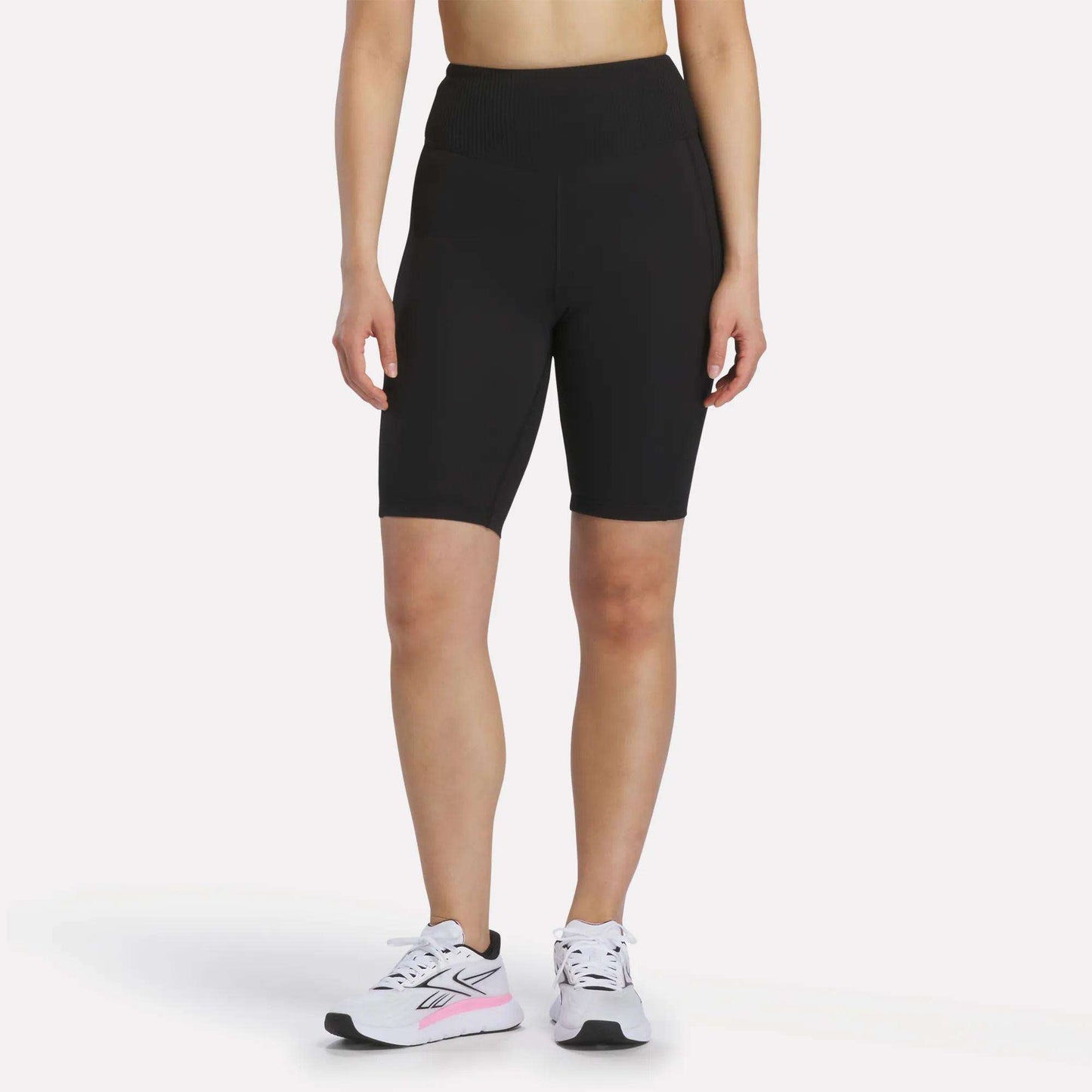 A person is shown from the shoulders down against a white background, wearing Reebok Women's Peached Biker Shorts 9" in black, paired with white athletic sneakers featuring pink accents.