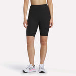 A person is shown from the shoulders down against a white background, wearing Reebok Women's Peached Biker Shorts 9" in black, paired with white athletic sneakers featuring pink accents.