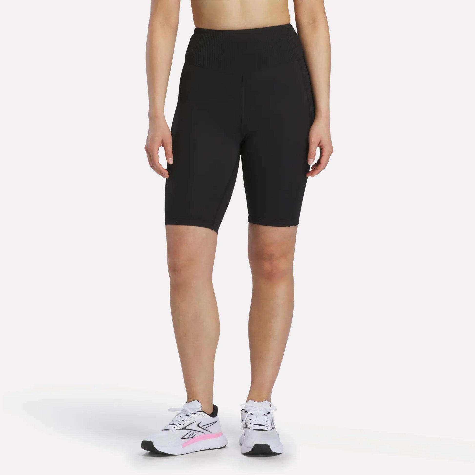 A person is shown from the shoulders down against a white background, wearing Reebok Women's Peached Biker Shorts 9" in black, paired with white athletic sneakers featuring pink accents.