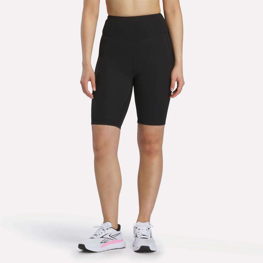 A person is shown from the shoulders down against a white background, wearing Reebok Women's Peached Biker Shorts 9" in black, paired with white athletic sneakers featuring pink accents.