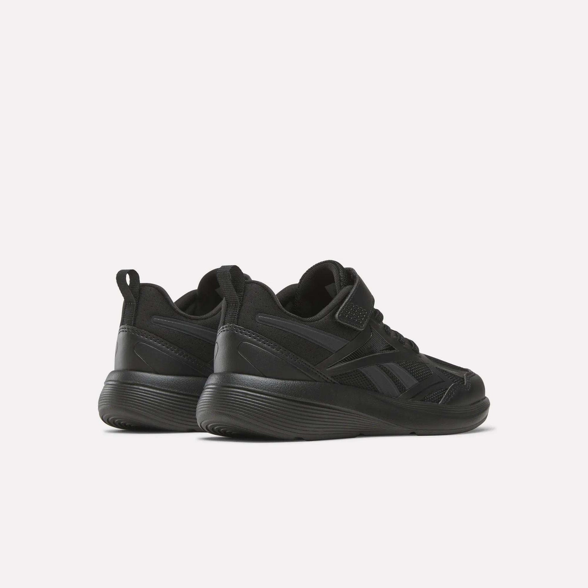 A pair of black Reebok Rush Runner 6.0 Elastic Lace Top Strap Running Shoes for little kids, shown from a rear-side angle on a light background. These low-top shoes have thick soles and subtle panel details, ideal for all-day play.