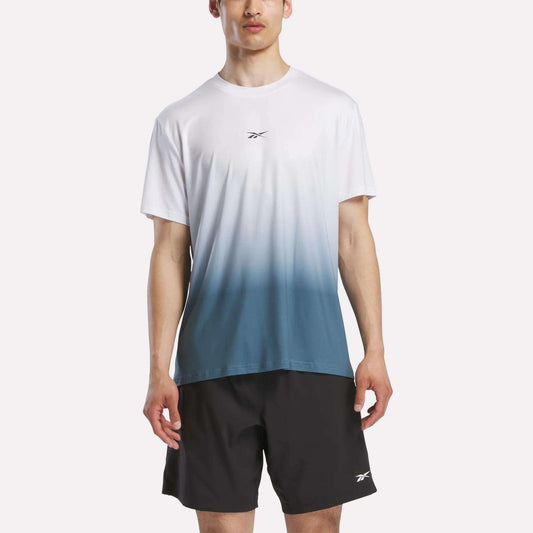 A man faces forward against a plain white background, wearing a Reebok Men's Athlete Speed Ombré T-Shirt in white and blue gradient, paired with black Reebok shorts.