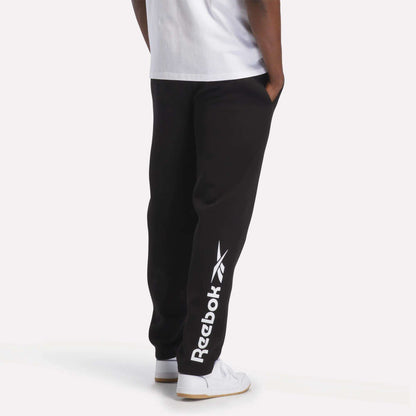 A man is wearing Reebok Men's Fleece Straight Leg Pants in black, featuring a white logo, paired with a white T-shirt and white sneakers. He stands with his hands in his pockets against a plain white background. These pants are crafted from cozy brushback fleece for all-day comfort and classic athletic style.