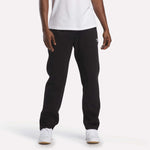 A person stands against a plain white background, with only the lower half of their body visible, showcasing the cozy comfort of the Reebok Men's Fleece Straight Leg Pants in black. They are styled here with a white t-shirt and white sneakers.