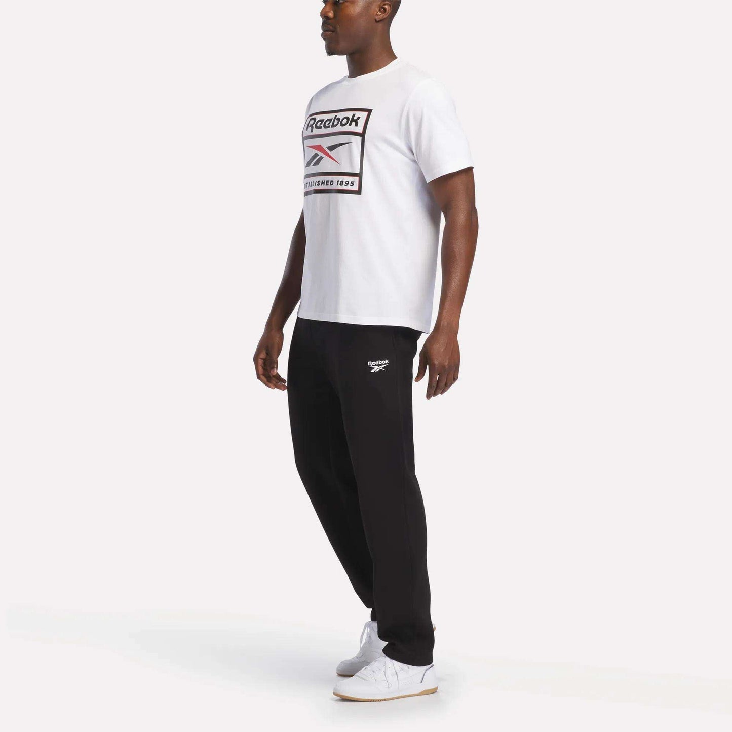 A man stands against a plain white background wearing a white Reebok t-shirt, Men's Fleece Straight Leg Pants from Reebok in black for cozy comfort, and white sneakers.