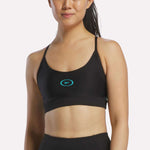 A woman wears the Reebok Women's Speed Strappy Sports Bra in Black/Twisted Turquoise, featuring thin straps and a small blue logo, standing against a plain light background.