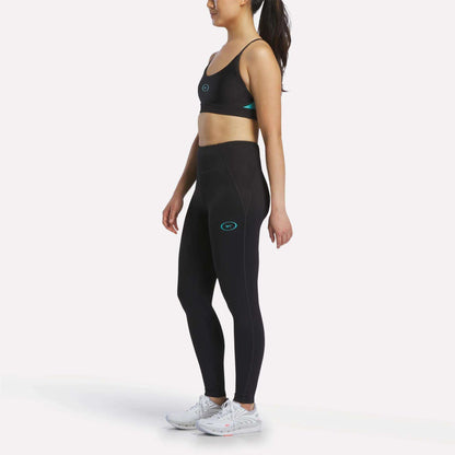 A woman poses in profile wearing the Reebok Women's Speed Strappy Sports Bra, black leggings with teal accents, and white athletic shoes against a plain light background.