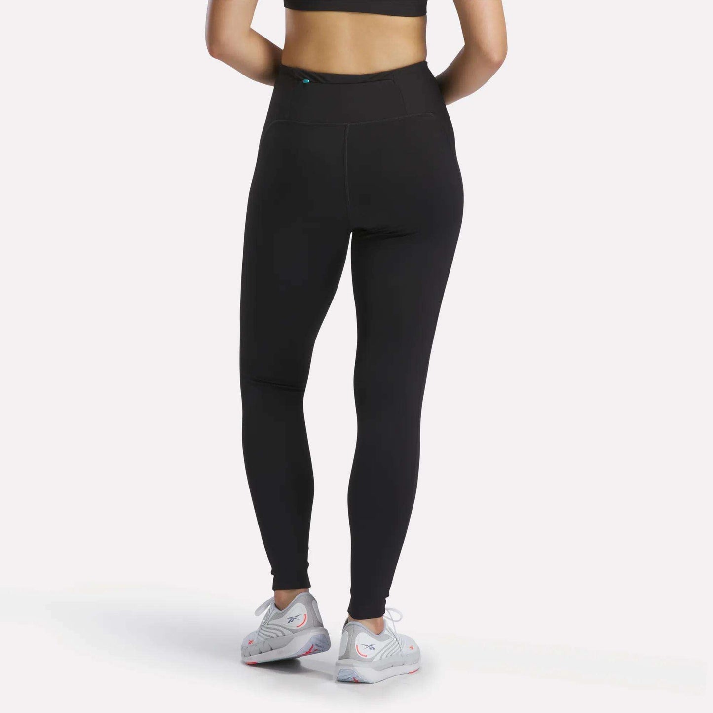 A person wearing Reebok Women's Speed HR High-Rise Leggings in black, a black sports bra, and white athletic shoes stands facing away from the camera against a plain white background.