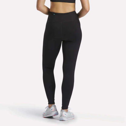 A person wearing Reebok Women's Speed HR High-Rise Leggings in black, a black sports bra, and white athletic shoes stands facing away from the camera against a plain white background.