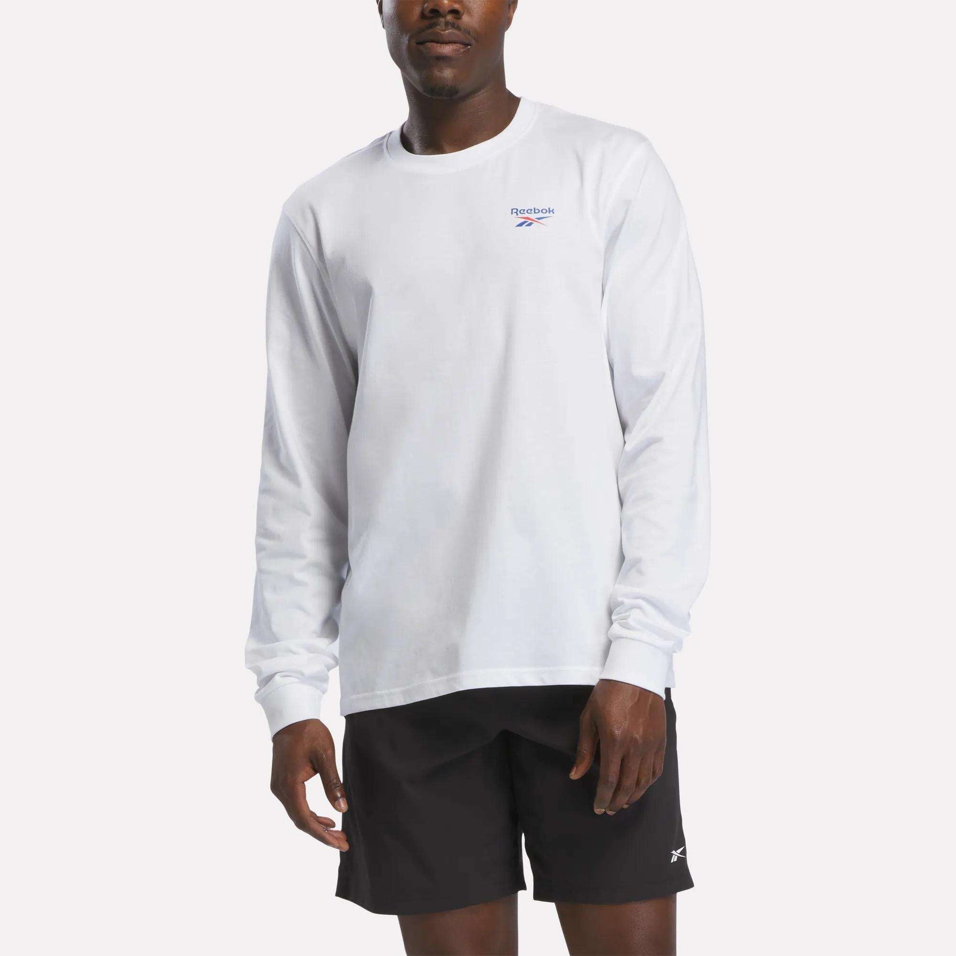 A person wearing a Reebok Men's Long Sleeve T-Shirt in white and black Reebok shorts stands against a plain, light background.