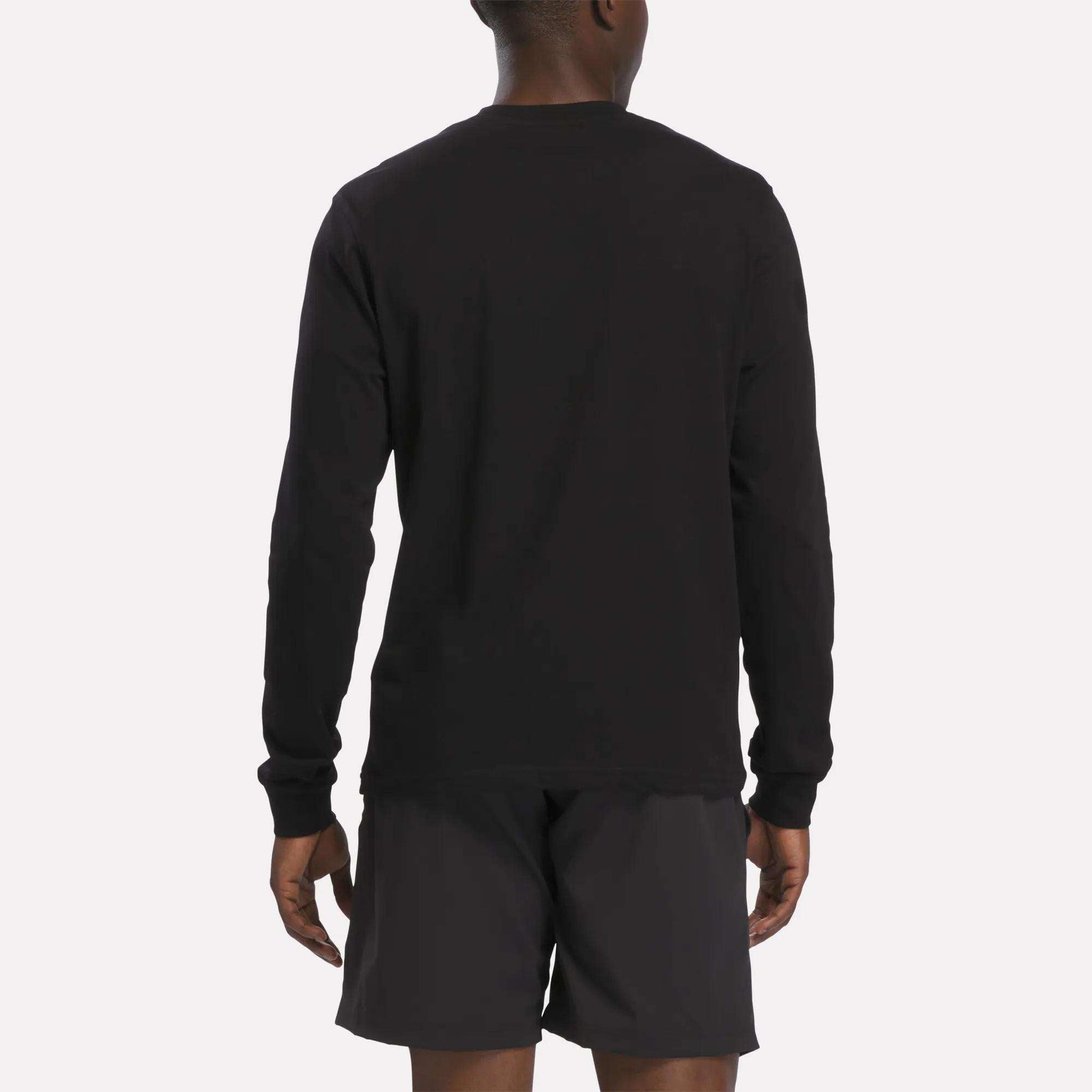 A person is standing facing away, wearing a Reebok Men's Long Sleeve T-Shirt in black and black shorts. The background is plain and light-colored.