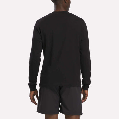 A person is standing facing away, wearing a Reebok Men's Long Sleeve T-Shirt in black and black shorts. The background is plain and light-colored.