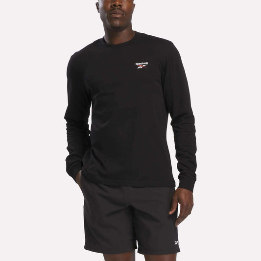 A person wearing a Reebok Men's Long Sleeve T-Shirt in black stands with one hand in their pocket against a plain light background.