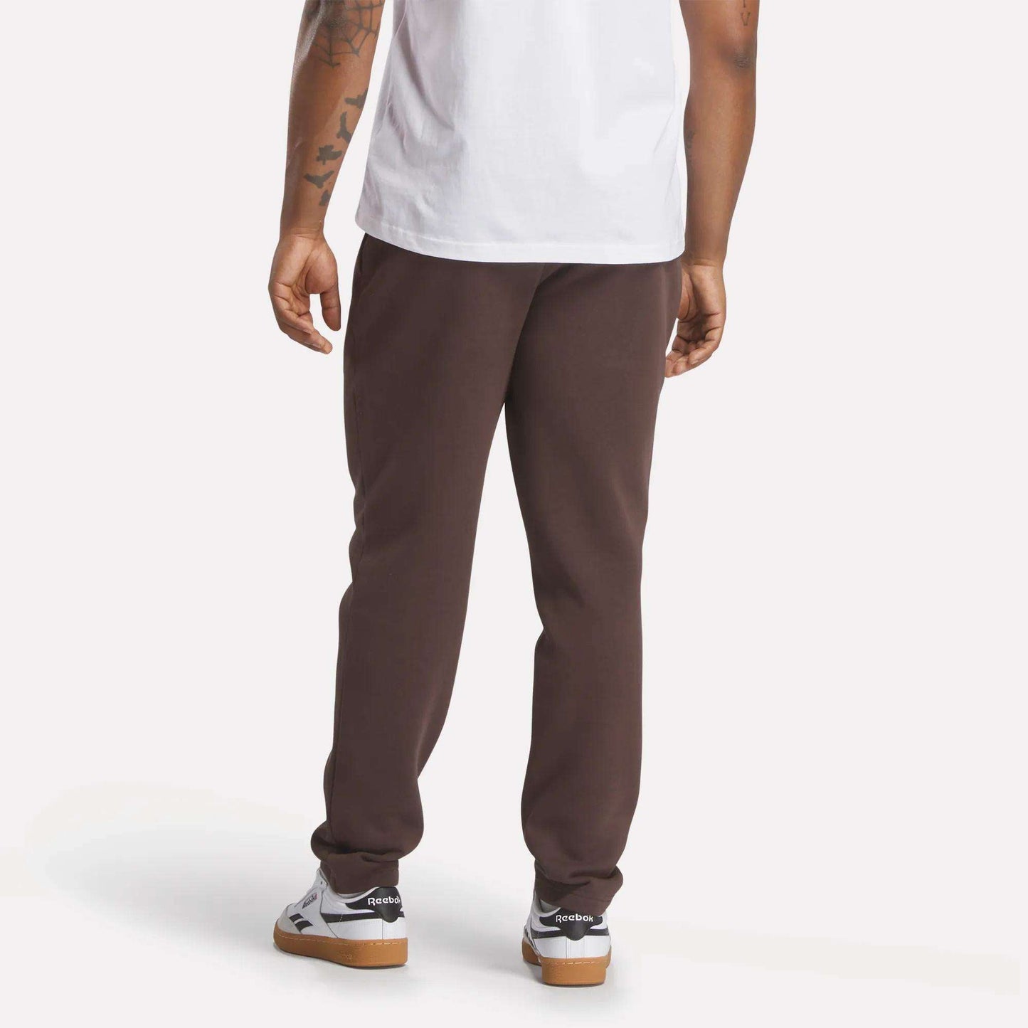 A person stands with their back to the camera, wearing a white t-shirt and slim fit dark brown Men's Patent Pants by Reebok, made from a cotton-poly blend, with white sneakers. Their tattooed right arm is visible against a plain light background.