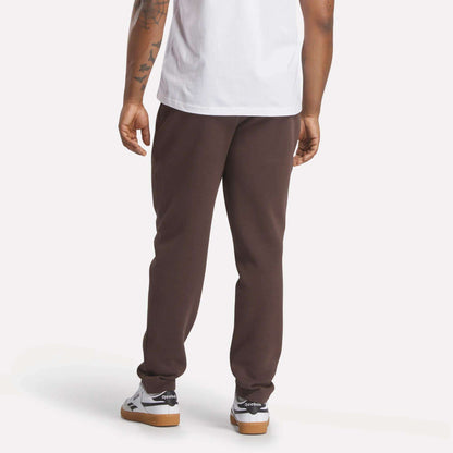 A person stands with their back to the camera, wearing a white t-shirt and slim fit dark brown Men's Patent Pants by Reebok, made from a cotton-poly blend, with white sneakers. Their tattooed right arm is visible against a plain light background.