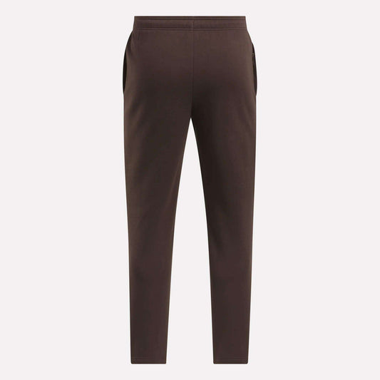 The Reebok Men's Patent Pants in brown are shown flat from the back on a light background, featuring an elastic waistband and side pockets. Made with a cotton-poly blend for comfort and durability.