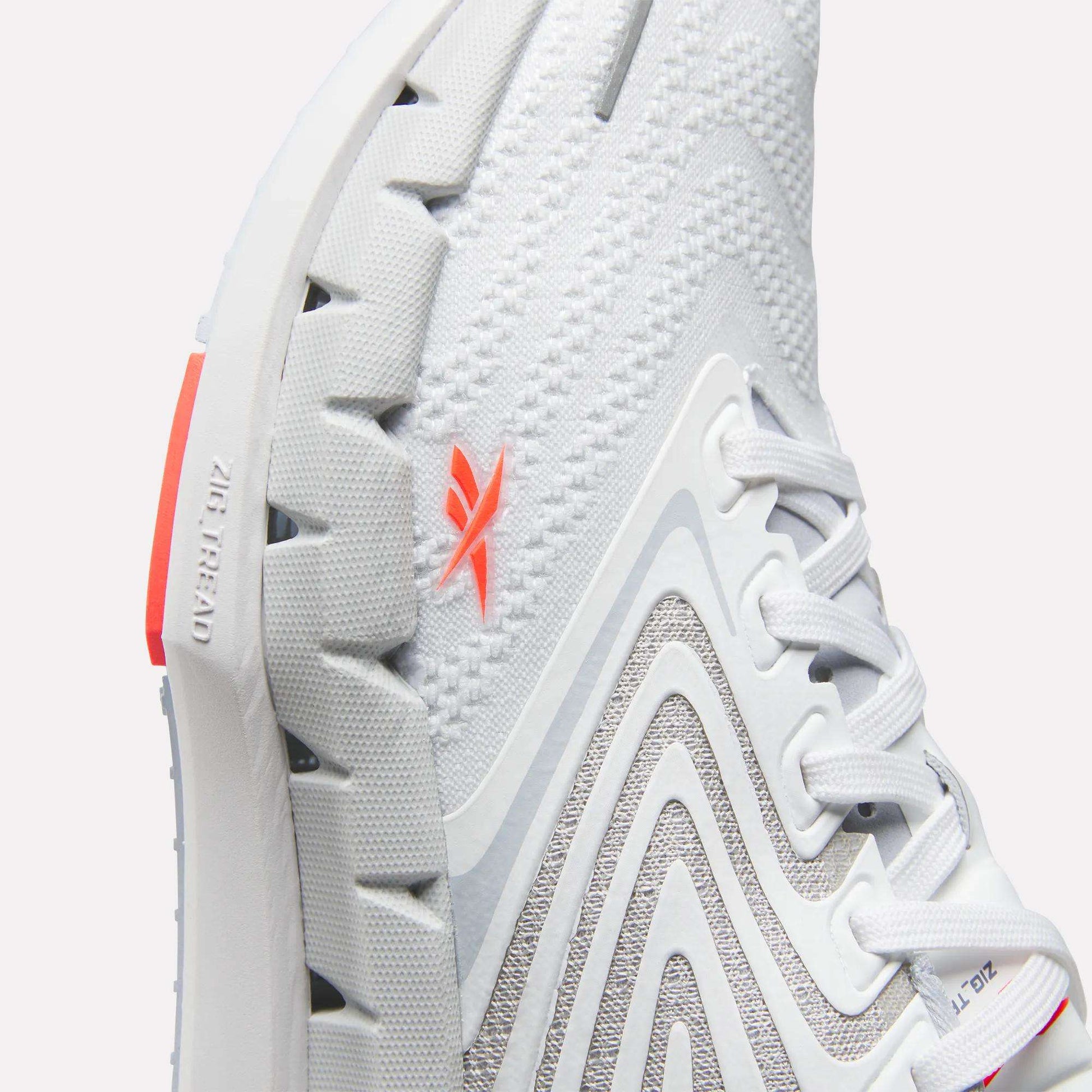 Close-up of Reebok Women's FloatZig Tread Running Shoe with breathable mesh, textured sole, gray curved patterns, white laces, and an orange logo. “Zig Kinetica” and “Zig” are embossed near the heel on the sole.