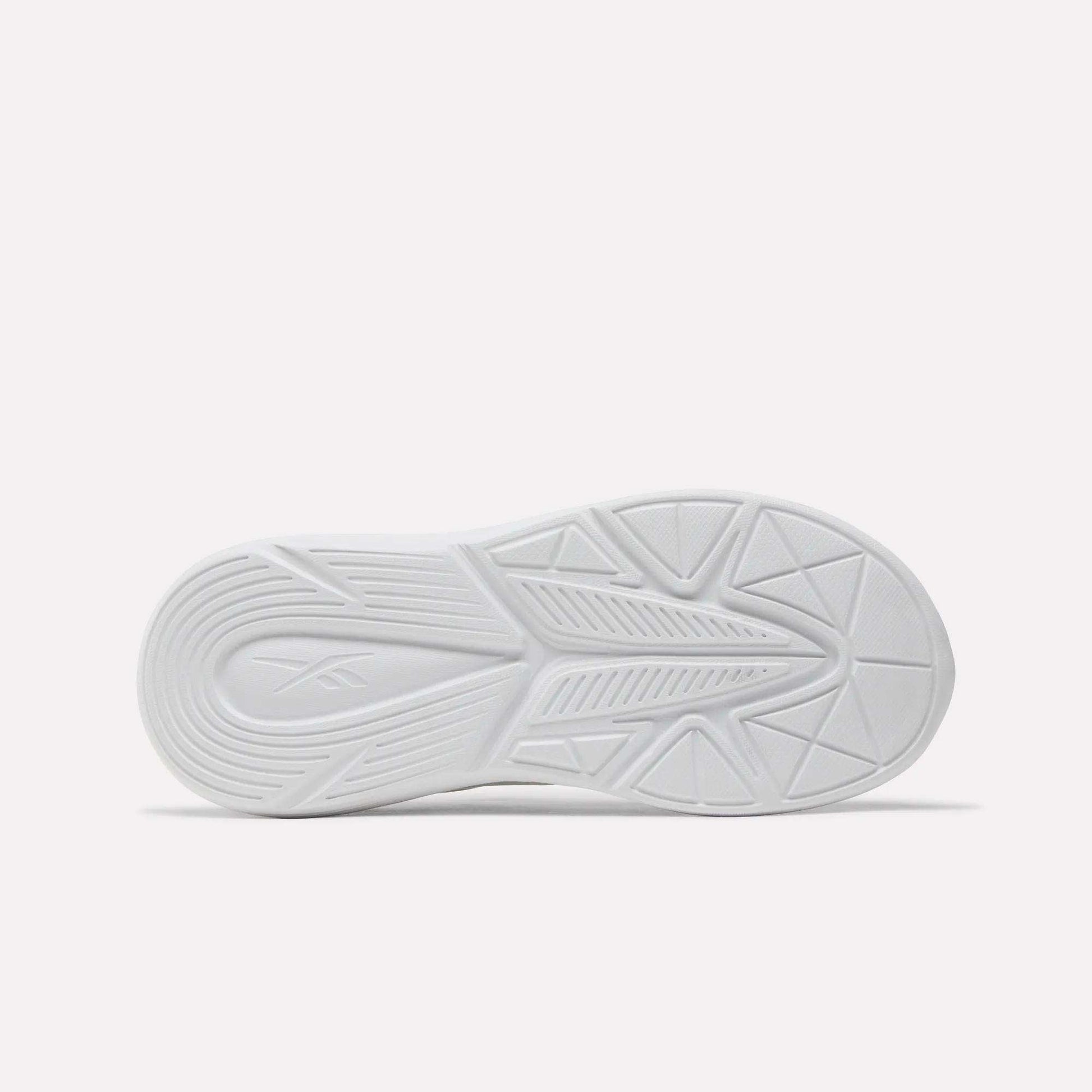 The image shows the bottom view of a white Reebok Rush Runner 6.0 Elastic Lace Top Strap Running Shoe for little kids, featuring geometric tread patterns and an embossed Reebok logo on the left, plus EVA cushioning for comfort. The background is plain and light-colored.