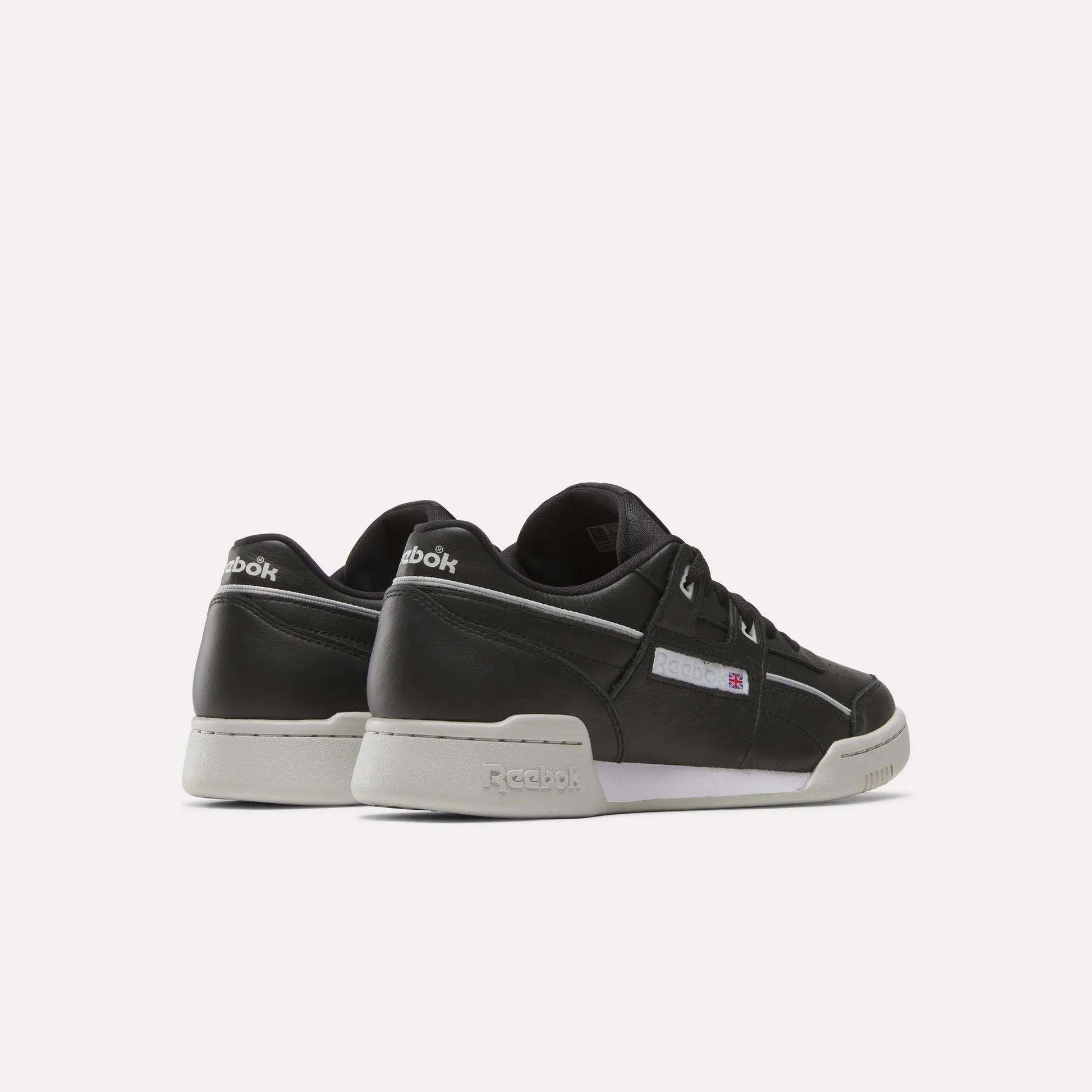 A pair of Reebok Workout Plus Shoes in black leather with white soles, displayed from the rear on a light background. The shoes feature Reebok branding on the heel, side, and tongue.