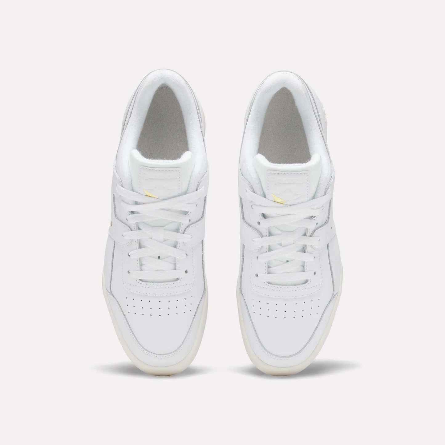 A pair of Reebok Workout Plus Shoes in white, featuring white laces, light yellow accents, perforated toe boxes, and cream outsoles, are seen from above on a plain background for an '80s-inspired look.