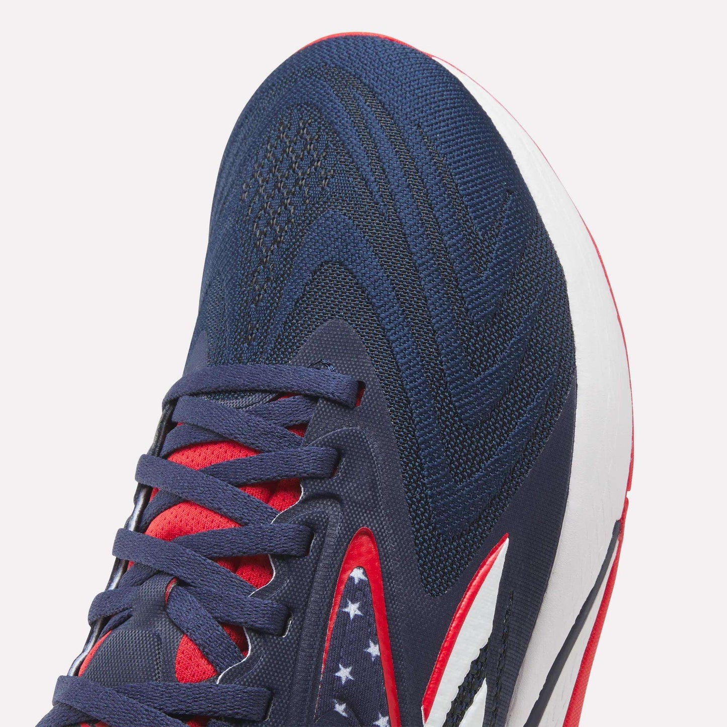 Close-up of Reebok Nano X5 Edge Training Shoes in navy with red and white accents, patterned laces, and white stars on a dark blue section. Featuring a Flexweave upper and white midsole for durability—ideal for precision training.