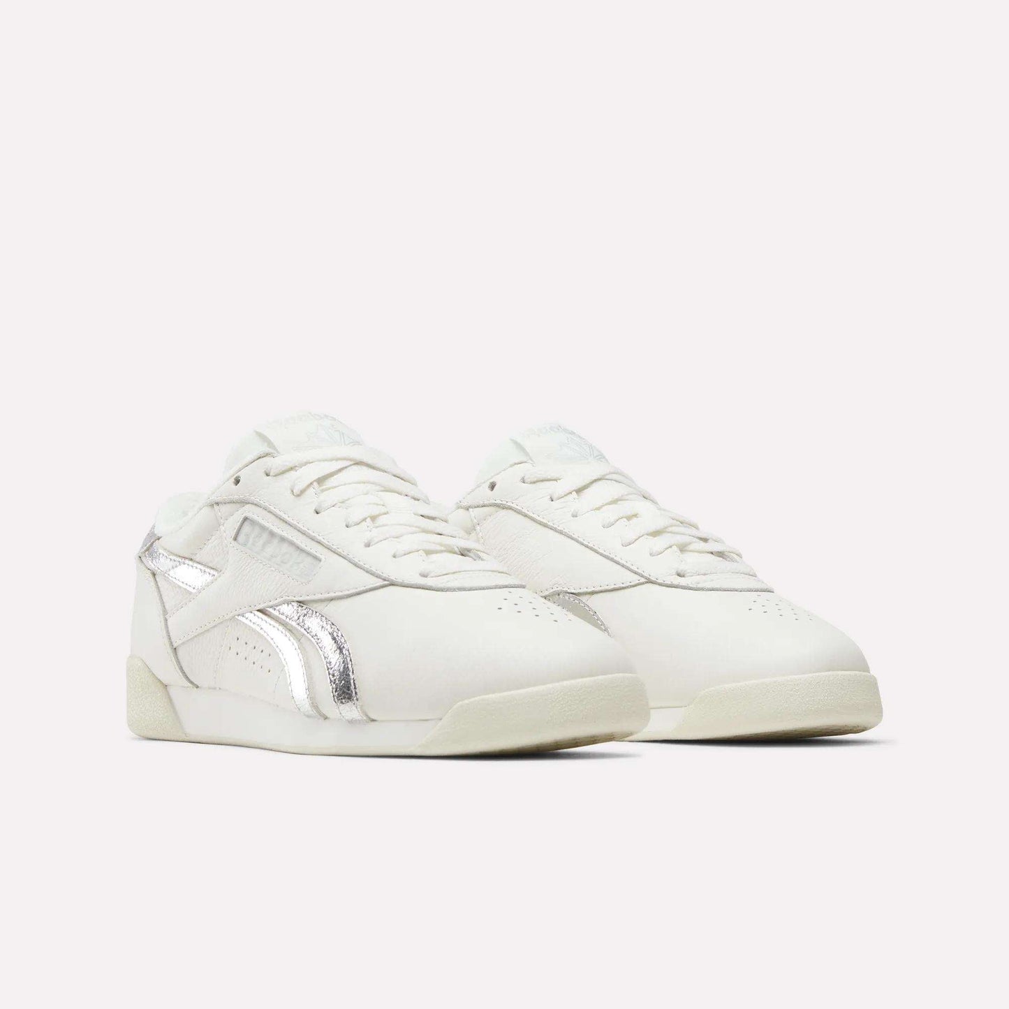 A pair of Reebok Women's Freestyle Lo Shoes with silver metallic accents is shown against a plain light background. These sneakers have a low-top design and sport a minimalistic style.