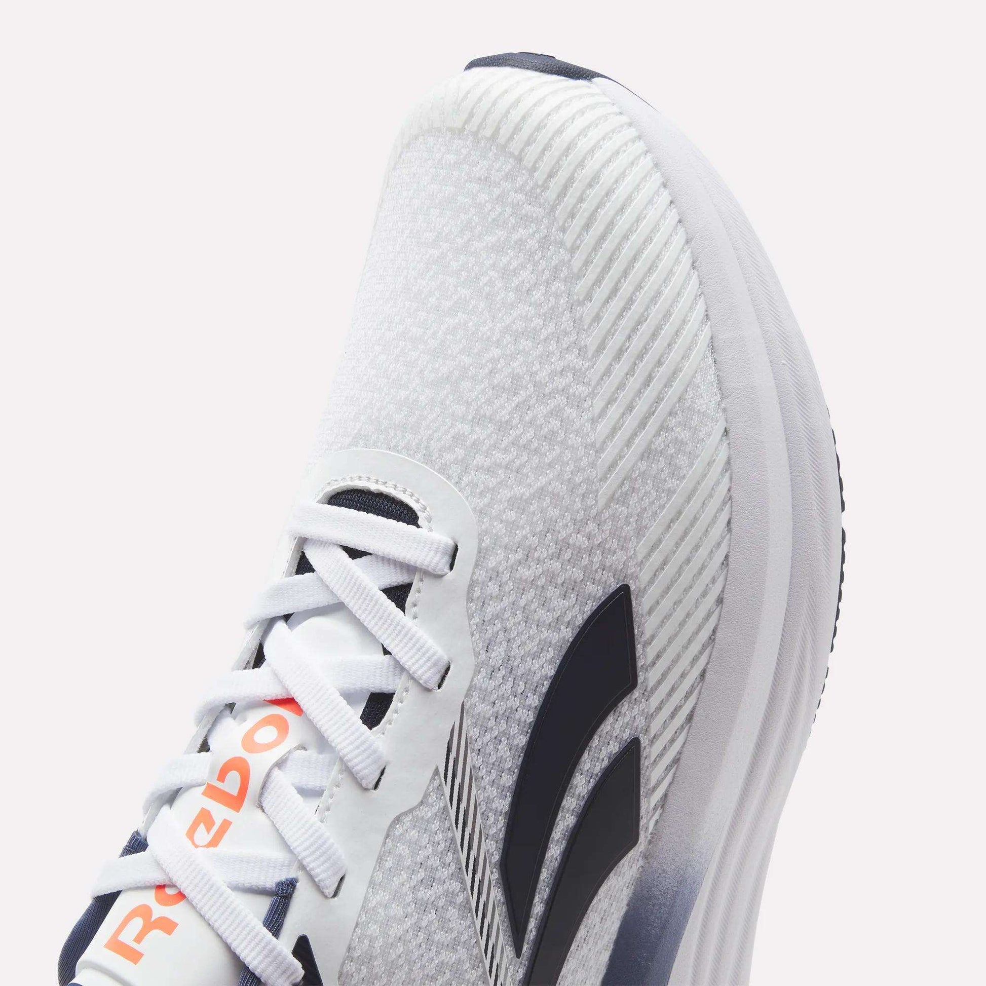 Close-up of a white Reebok Men's Viva Speed Shoe featuring black stripes, a gray knit texture, and orange Reebok branding on the tongue. Designed for ultimate comfort, these shoes include white laces, a thick cushioned sole, and a breathable knit construction—making them ideal for active lifestyles.