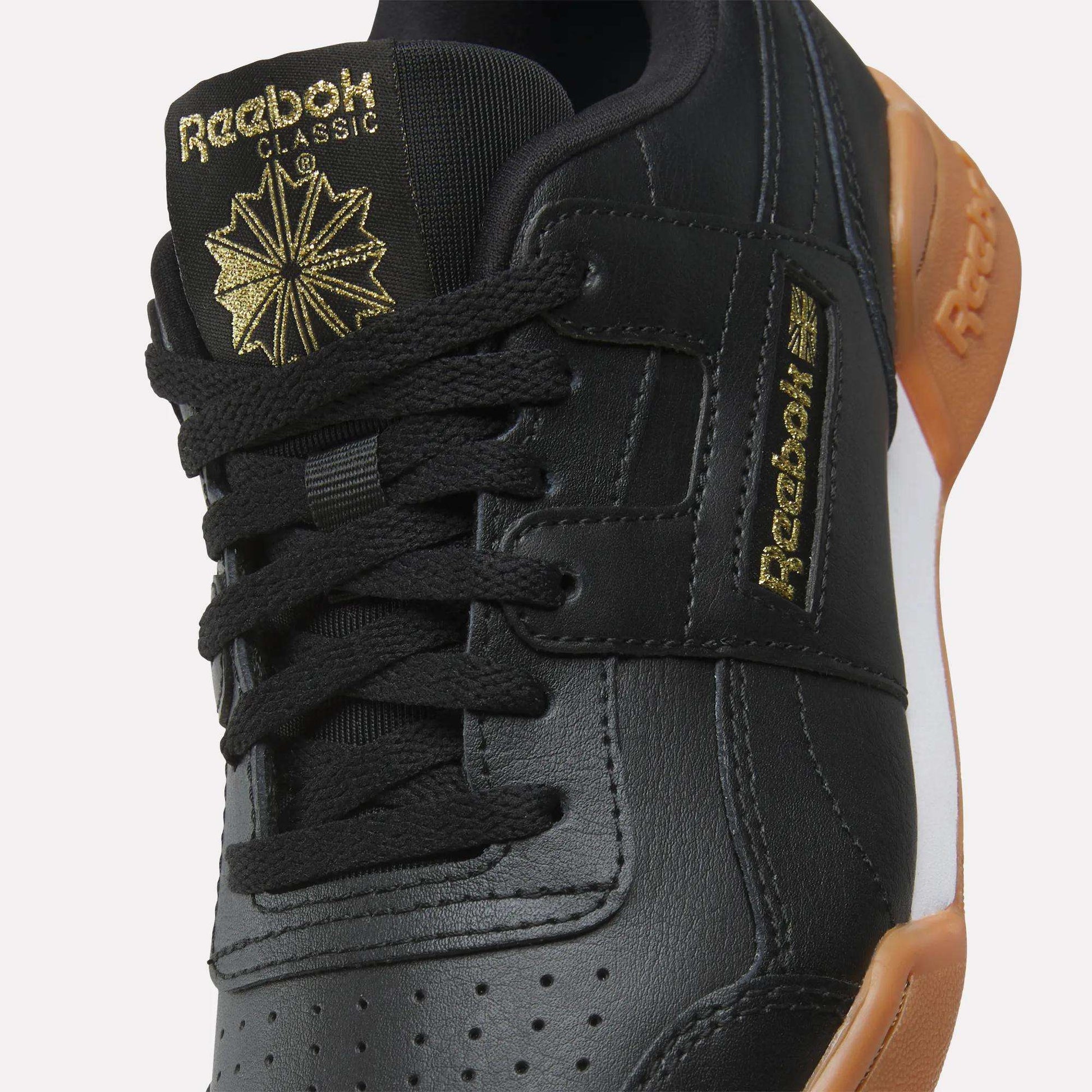Close-up of black Reebok Workout Plus Shoes for big kids, featuring black laces, gold Reebok branding on the tongue and side, and a brown rubber sole—an eye-catching choice among Reebok footwear.
