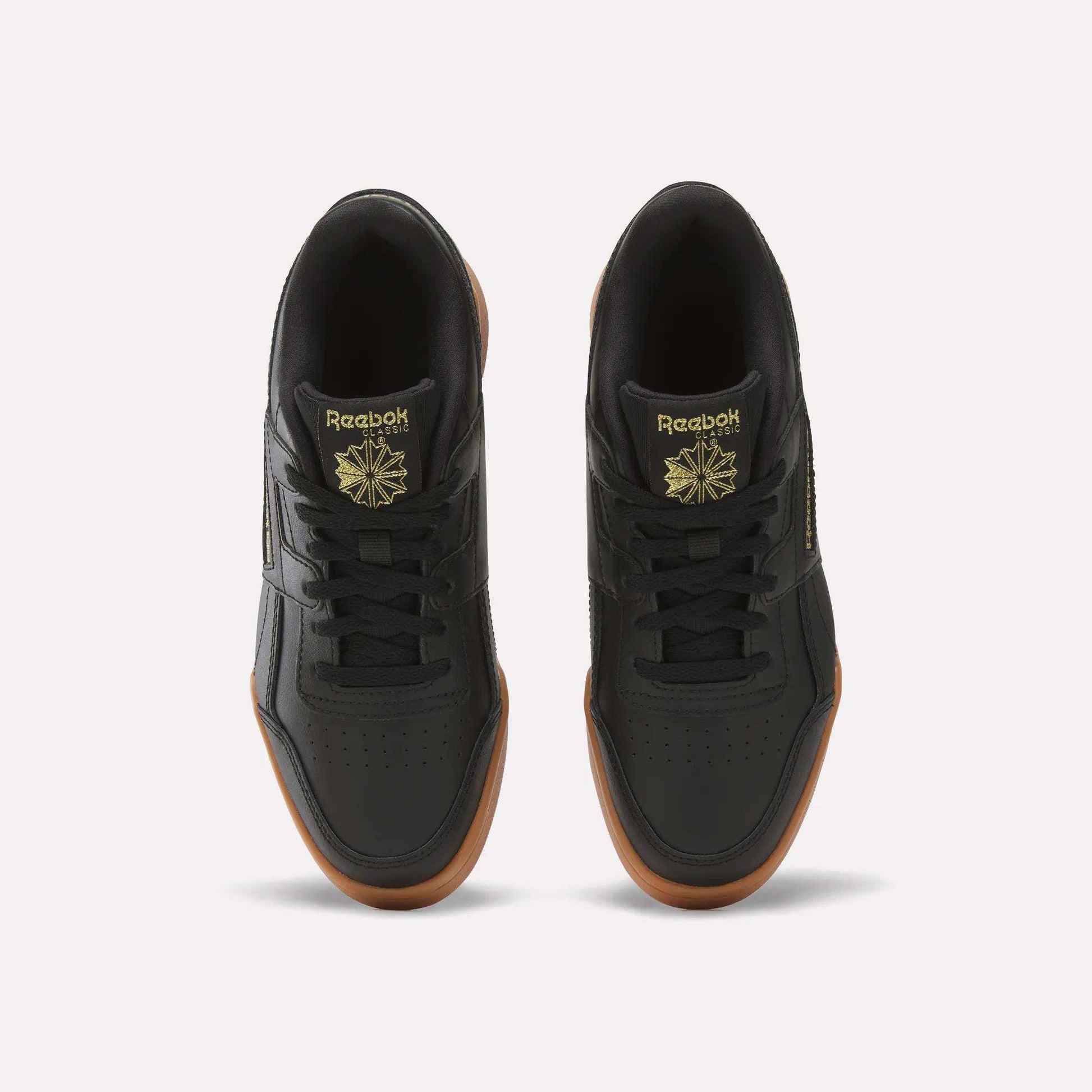 Top view of Reebok Workout Plus Shoes for big kids in black with brown gum soles, gold branding, and a star logo on the tongue, side by side on a white background—a sporty pick for juniors.
