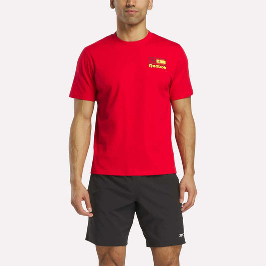 A man stands against a white background in a bright red Reebok España Football T-Shirt with a small flag and logo on the chest, paired with black Reebok shorts.