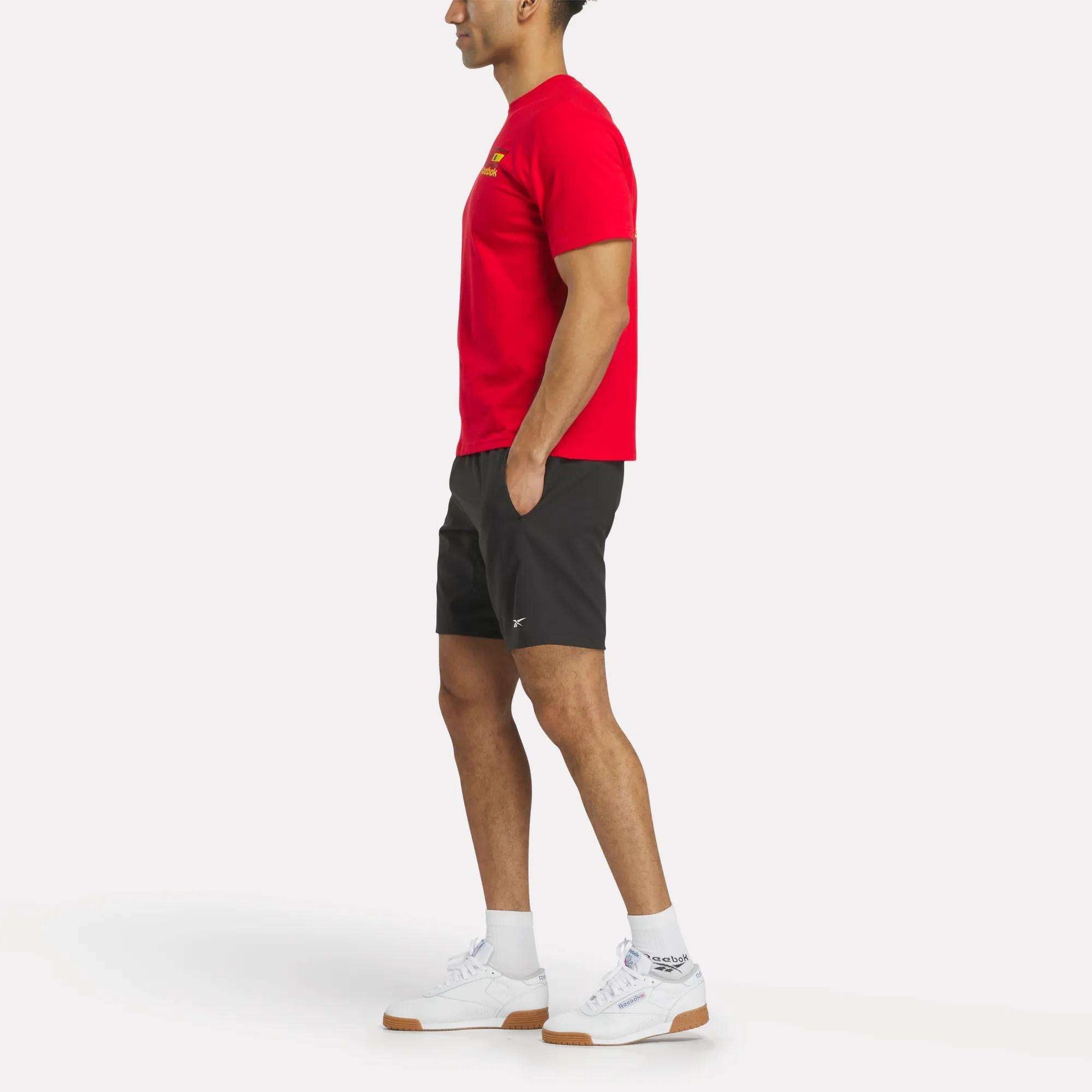 A person stands in profile against a plain white background, wearing an España Football T-Shirt and white Reebok sneakers, paired with black shorts and white socks.