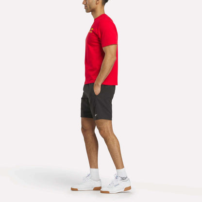 A person stands in profile against a plain white background, wearing an España Football T-Shirt and white Reebok sneakers, paired with black shorts and white socks.