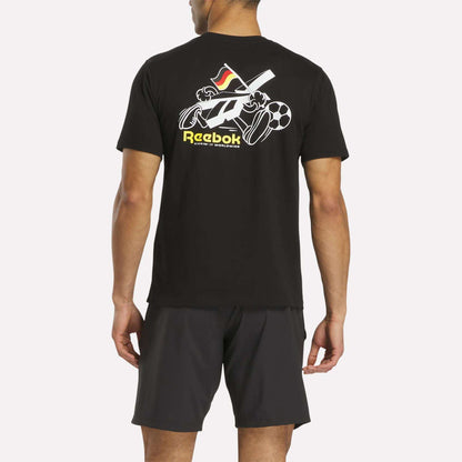 A man stands facing away, wearing a black Reebok Deutschland Football T-Shirt featuring sports equipment graphics and Reebok text on the back, paired with dark shorts.