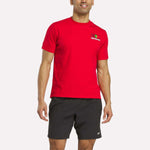 A man wears a bright red Reebok Portugal Football T-Shirt with a small logo on the left chest and black athletic shorts with bold team graphics, standing against a plain white background.