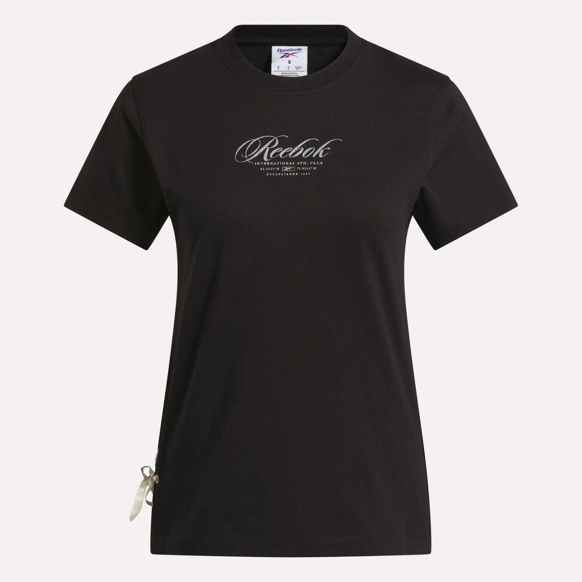 The Reebok Women's Ribbon T-Shirt is shown in black with short sleeves against a white background. It features the brand’s cursive logo on the chest and a small ribbon detail at the bottom left hem. Made from soft cotton, this t-shirt offers comfortable wear and metallic shine for a stylish everyday look.